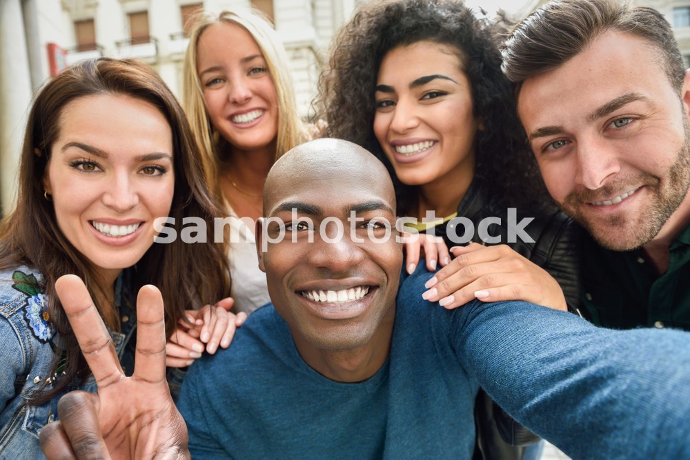 Multiracial group of friends taking selfie in a urban street with a black man in foreground. Three young women and two men wearing casual clothes.