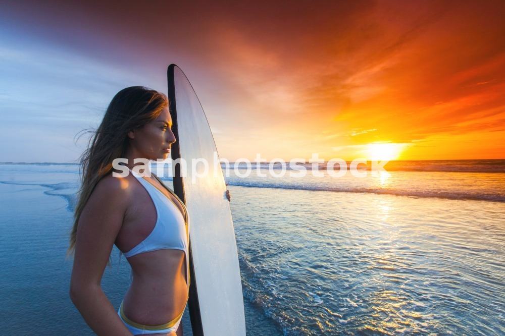 A beautiful young slim sporty woman in bikini with a surfboard is standing at ocean beach at sunset. Woman with surfboard