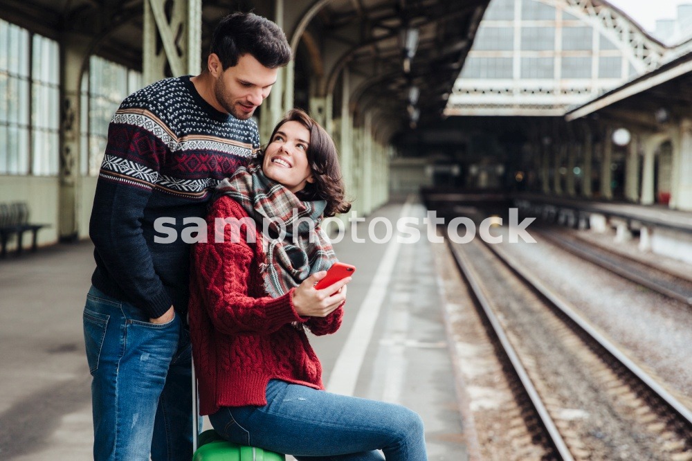 Family couple being on platform, wait for train. Beautiful woman sits on suitcase, uses smart phone, looks at her lover, shows him photos. Young woman and man use cell phone on railway station